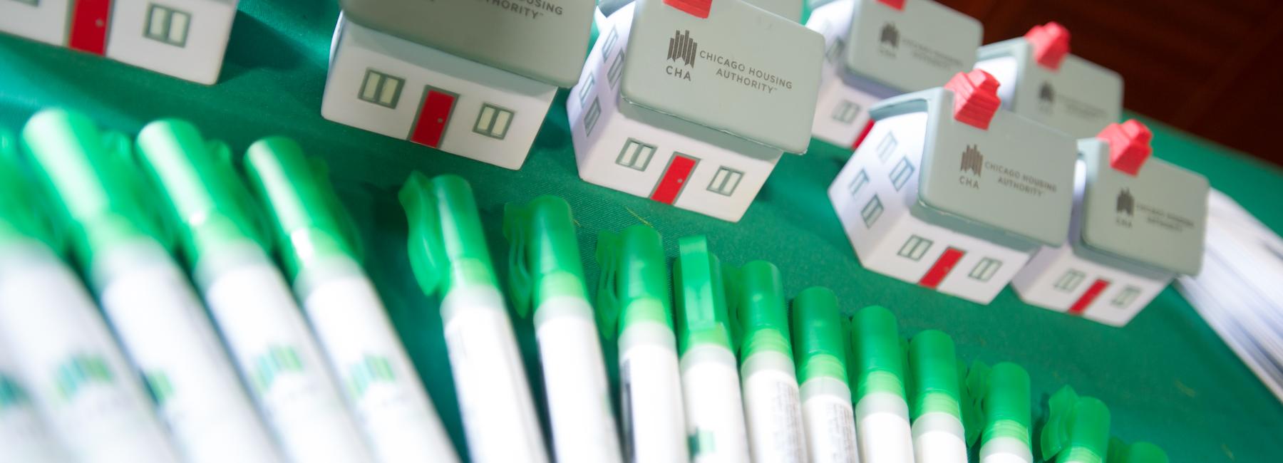 The image shows a table with several miniature house models and small spray bottles lined up. The house models and mini hand sanitizer spray bottles both have the logo of the Chicago Housing Authority (CHA) printed on them. The house models feature grey roofs with red chimneys, white walls with green window frames, and red doors. The hand sanitizer spray bottles have green caps. The background is blurred but appears to be a green tablecloth covering the table.