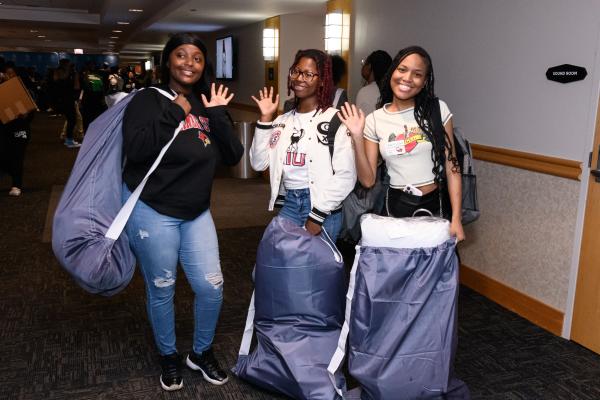 Three young women smile and wave at the camera while holding large purple bags. They are standing in a hallway with other people in the background. One woman wears a black sweatshirt and ripped jeans, the second woman wears a white letterman jacket and jeans, and the third woman wears a light-colored t-shirt and dark jeans.