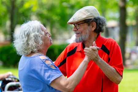 An older man wearing a golf hat and an orange shirt dances with a gray-haired woman in a blue shirt.