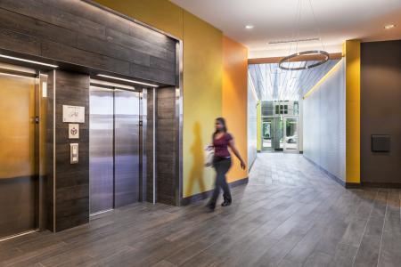 A person walks through a modern, well-lit hallway with yellow and gray walls, towards a set of stainless steel elevators. The hallway has dark wood flooring and a large circular light fixture hanging from the ceiling. A glass door entrance is visible in the background.