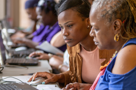 Two women are sitting closely together, focused on a task at a computer. The younger woman, with short hair, is using a mouse and looking intently at the screen, while the older woman, with braided hair and wearing a blue top, watches closely. In the background, several other people are also working on computers, creating a busy, collaborative atmosphere. The scene appears to be a learning environment, such as a computer lab or resource center, with the two women sharing knowledge or working together on a p