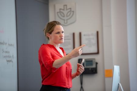 A woman wearing a red blouse is standing and gesturing with her right hand as if pointing at something. She is holding a whiteboard marker in her left hand. The setting appears to be a classroom or lecture room, with a whiteboard partially visible on the left and a wall with plaques or awards in the background. Her expression is focused and engaged, indicating she is likely teaching or giving a presentation.