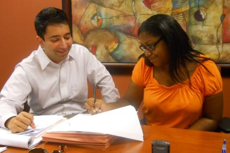 A man and a woman are seated at a table, engaged in a collaborative activity, likely reviewing or signing documents. The man, dressed in a light striped shirt, is smiling and looking down at the papers. The woman, wearing glasses and an orange top, is holding a pen and appears to be writing or signing something. They are seated in a well-lit room with a large, abstract painting hanging on the wall behind them. The atmosphere seems professional yet friendly. Various office items, including a phone and a pair