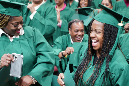 A group of joyful graduates wearing green caps and gowns celebrate together. One woman in the foreground is holding a certificate and laughing, while another next to her is holding a phone, capturing the moment. The room is filled with other graduates who are also smiling and cheering, creating a lively and festive atmosphere.