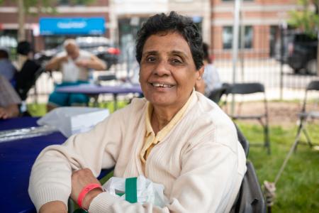 A smiling older woman is sitting at an outdoor event. She is wearing a beige sweater and a yellow shirt, with a red wristband on her left wrist. The woman is seated at a table with a blue tablecloth, and there are other people and tables in the background. She looks content and happy, with short curly hair and holding a white and green bag on her lap. The background includes a blurred view of buildings and greenery, indicating a community or festival setting.