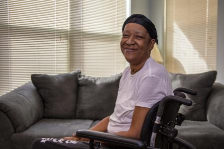 A smiling person wearing a white t-shirt and a black headscarf sits in a wheelchair in a room with a gray couch and window blinds in the background.