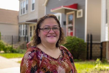 A woman with shoulder-length curly brown hair and glasses is smiling warmly at the camera. She is wearing a colorful blouse with red, black, and beige patterns. The background shows a residential neighborhood with houses, greenery, and a sunny, clear sky. The overall atmosphere is bright and welcoming.